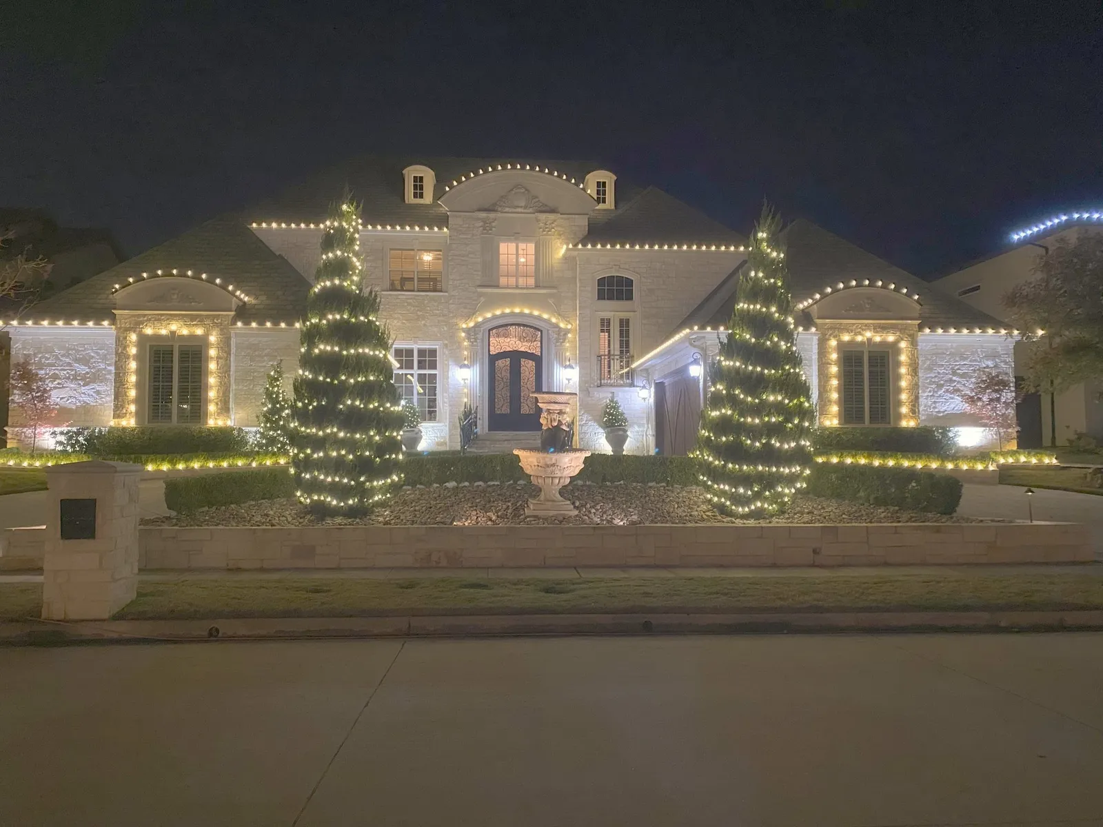 A large North Texas home outlined in warm white Christmas lights with two tall evergreens wrapped trunk to tip
