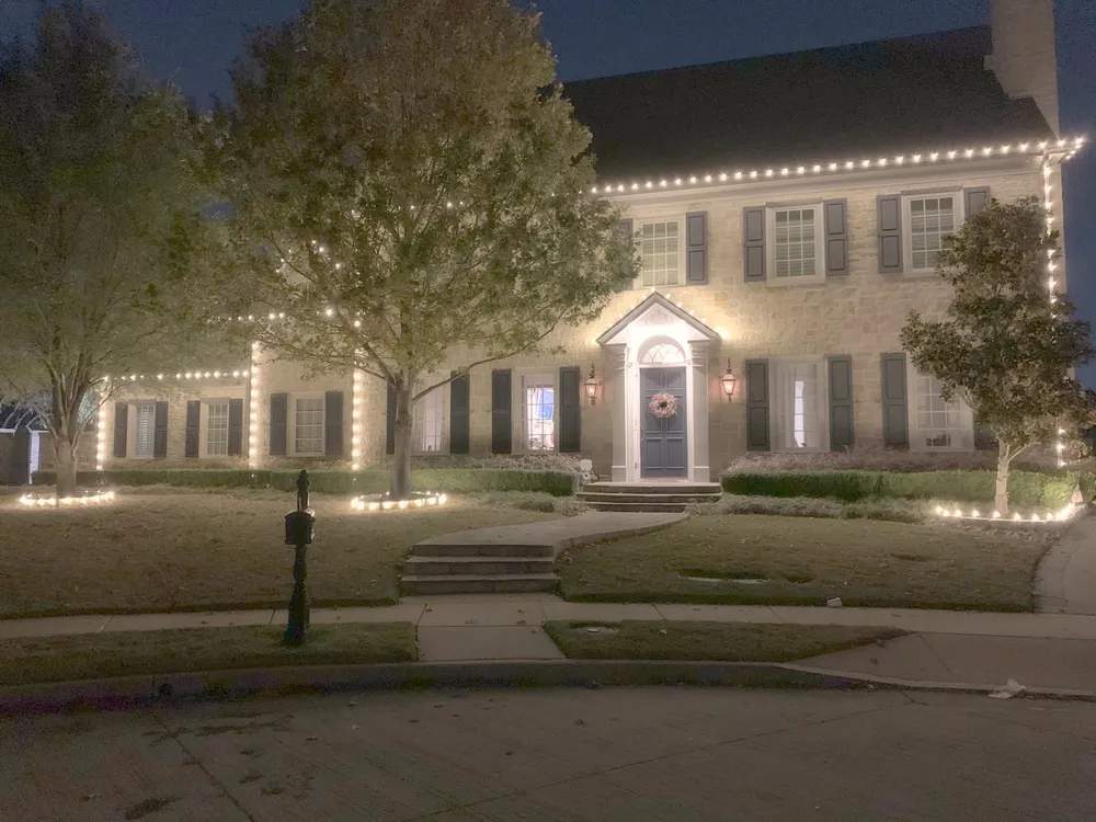 A colonial home lit in warm white with wrapped trees at night