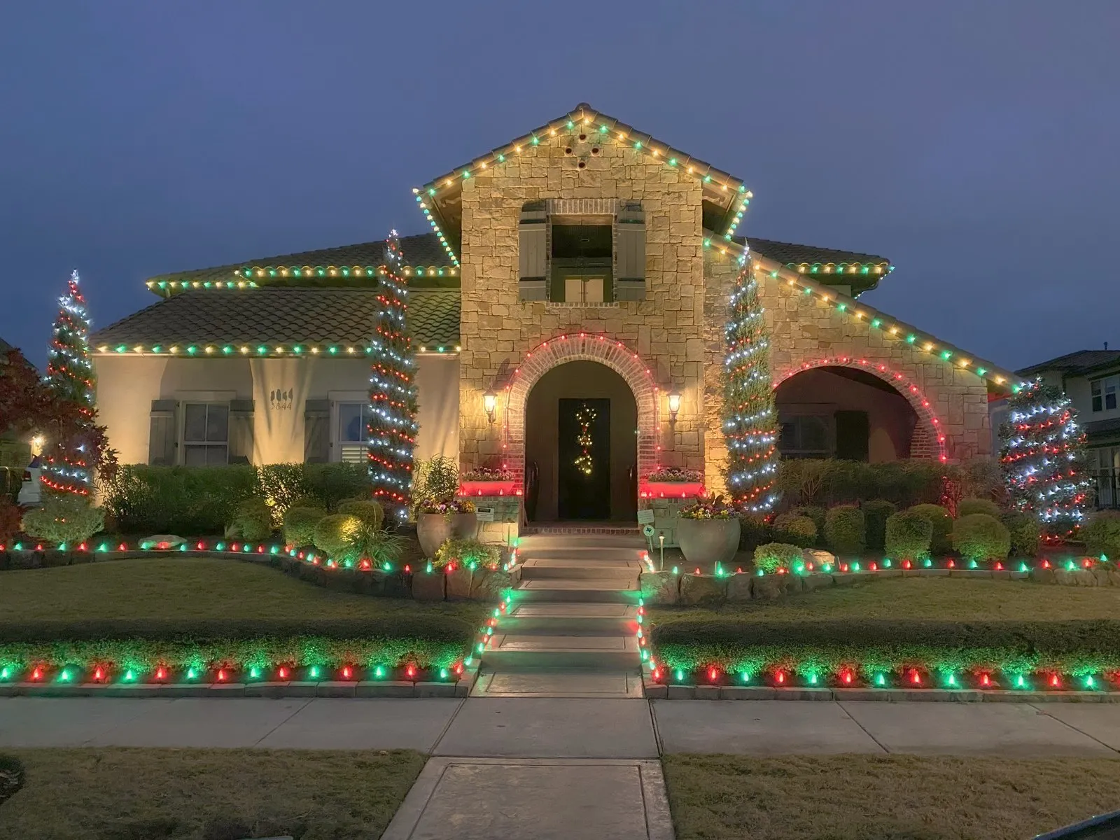 A North Texas home lit in red and green Christmas lights on the roofline, trees, and walkway at blue hour