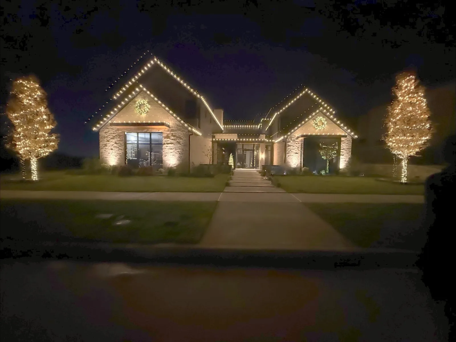 A ranch home at dusk flanked by two fully lit Christmas trees, installed by South Custom Lighting near Aubrey, Texas